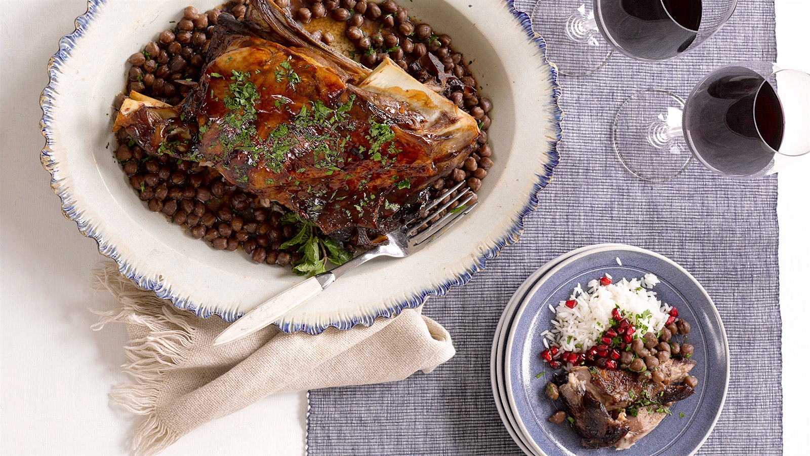  A platter of braised lamb shoulder and chickpeas; a plate with the lamb, rice and pomegranate seed garnish; and two glasses of red wine